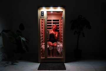 A woman sits inside an illuminated Lifepro infrared sauna cabin in a dark room. The sauna’s red lights glow brightly.