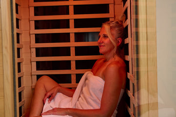 A woman wrapped in a white towel relaxes while leaning back inside a wooden home infrared sauna.