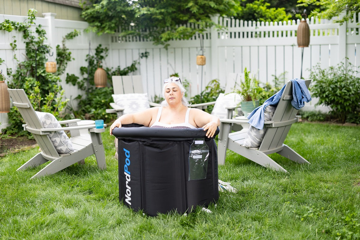 A woman resting with eyes closed inside a NordPod cold plunge ice tub setup on a backyard lawn.
